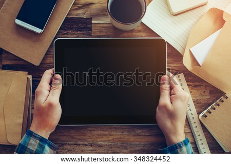 Examining his new tablet. Close-up top view of male hands holding digital tablet over wooden desk with different chancellery stuff laying on it