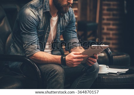 In search of new ideas. Close-up of young bearded man holding digital tablet and sitting in chair