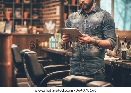 Digital technologies in any business. Close-up of young bearded man holding digital tablet while standing at barbershop
