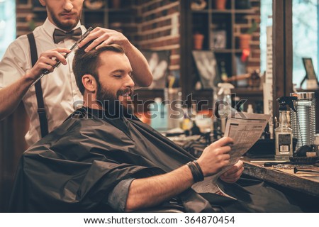 Great time at barbershop. Cheerful young bearded man getting haircut by hairdresser and reading newspaper while sitting in chair at barbershop