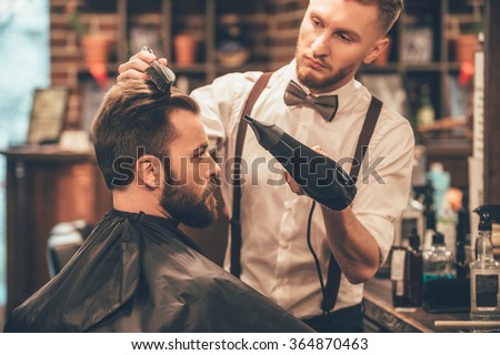 New hairstyle. Side view of young bearded man getting groomed at hairdresser with hair dryer while sitting in chair at barbershop
