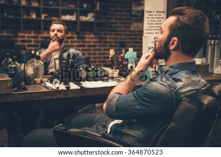 Looking good. Side view of handsome young bearded man looking at his reflection in the mirror and keeping hand on chin while sitting in chair at barbershop