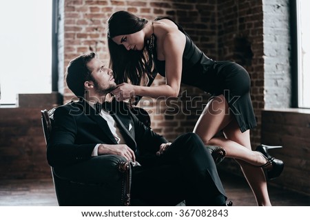 Time to seduce. Beautiful young woman in cocktail dress leaning to her boyfriend sitting in chair while looking at each other in loft interior