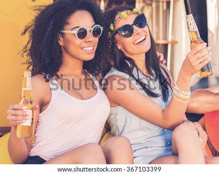Enjoying carefree time. Two cheerful young women holding bottles with beer and smiling while while sitting outdoors together