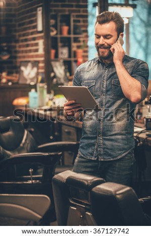 our appointment is on Monday! Cheerful young bearded man talking on mobile phone and looking at digital tablet while standing at barbershop