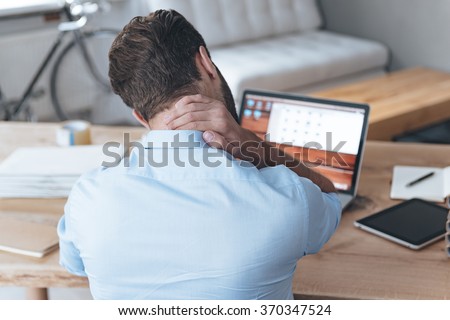 Feeling exhausted. Rear view of frustrated young man looking exhausted and massaging his neck while sitting at his working place