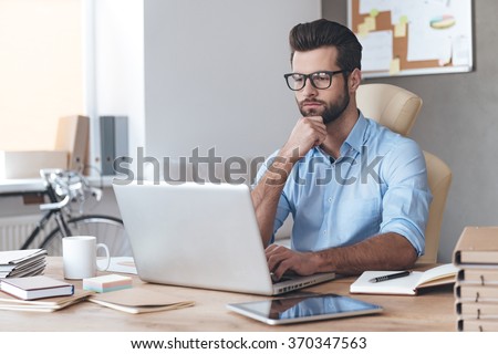 Busy working. Pensive young handsome man wearing glasses working on laptop and keeping hand on chin while sitting at his working place
