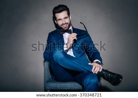 Well hello! Cheerful young handsome man in suit holding his sunglasses and looking at camera with smile while sitting on the chair against grey background