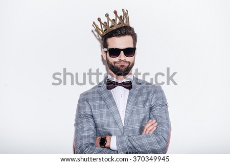 King of style. Sneering young handsome man wearing suit and crown keeping arms crossed and looking at camera while standing against white background