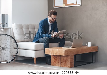 Multitasking. Handsome young man wearing glasses and working with touchpad while sitting on the couch in office