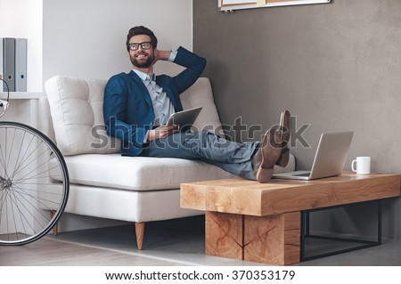 Enjoying his working day. Handsome cheerful young man keeping legs on table and looking away with smile while sitting on the couch in office