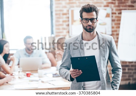 Confident businessman. Young handsome man holding notepad and looking at camera while his colleagues discussing something in the background