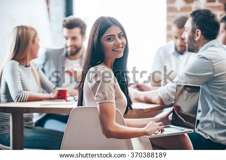 Glad to be a successful businesswoman. Cheerful young woman holding laptop on her knees and looking at camera while her colleagues discussing something in the background