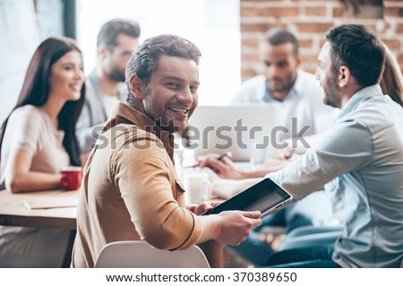 Glad to be a part of team. Cheerful young man holding digital tablet and looking at camera while his colleagues discussing something in the background
