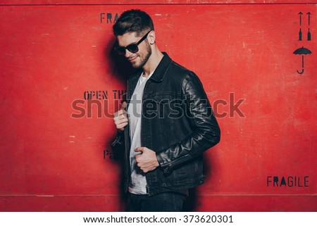 Trendy look. Handsome young man in sunglasses adjusting his jacket and looking away with smile while standing against red background