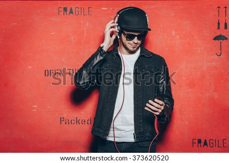 Style and music. Handsome young man adjusting his headphones and looking at his smart phone while standing against red background
