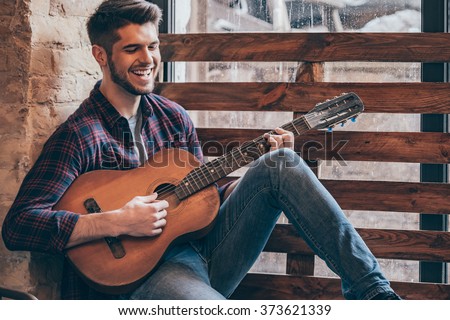 Cheerful guitarist. Cheerful handsome young man playing guitar and smiling while sitting at windowsill