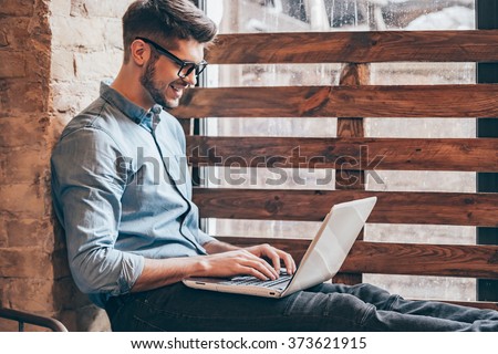 Blogger at work.Side view of handsome young man working on laptop and smiling while sitting at windowsill