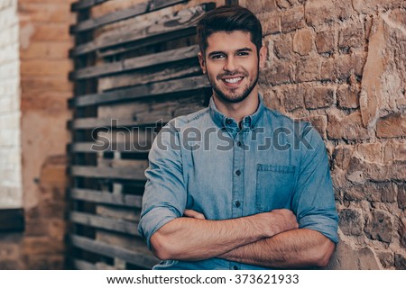 Smiling and handsome.Handsome young man keeping arms crossed and looking at camera with smile while standing against brick wall