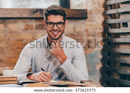 Working with pleasure. Handsome young man in glasses making some notes in his note pad and looking at camera with smile while sitting at his working place