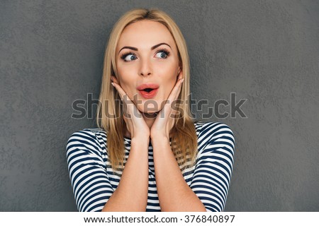 Oh! Beautiful young woman keeping hands on chin and looking surprised while standing against grey background