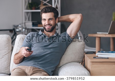 Great TV show.Handsome cheerful young man holding remote control and watching TV while sitting on the couch at home