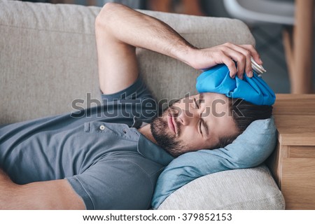 When you hit shelf with your head. Frustrated young man holding ice bag on his head while lying on the couch at home