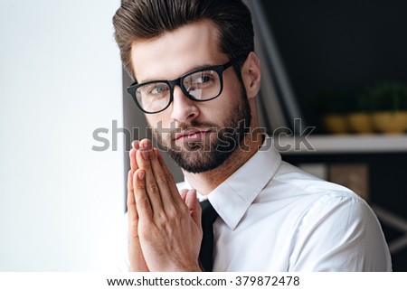 Calm and handsome. Handsome young businessman in glasses looking away and keeping hands clasped while standing in office