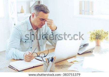 Feeling sick and tired. Frustrated mature man looking exhausted while sitting at his working place and carrying his glasses in hand