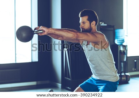 Intensive cross training. Side view of young handsome man in sportswear working out with kettle bell at gym