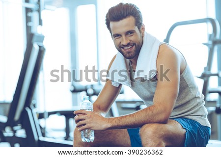 Little break. Handsome young men in sportswear holding water bottle and looking at camera with smile while sitting at gym