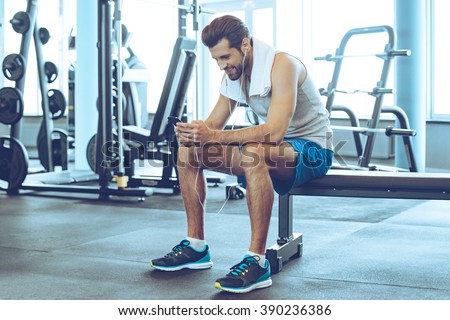 Satisfied with his work out. Handsome young men in headphones using his smart phone with smile while sitting at gym