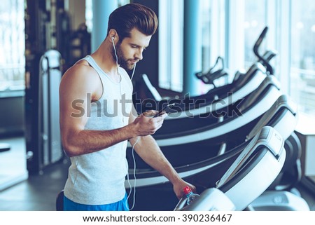 Best song for his training. Side view of young handsome man in sportswear using his smart phone while standing on treadmill at gym