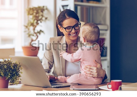 Working together is so fun! Cheerful young beautiful businesswoman looking at her baby girl with smile while sitting at her working place