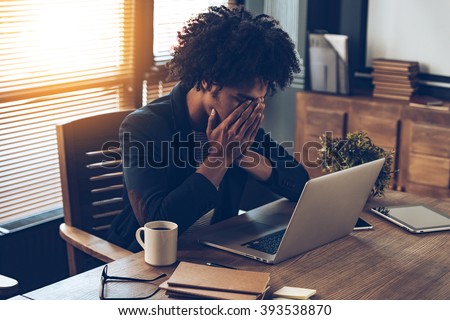 Total disappointment. Young African man looking exhausted and covering his face with hands while sitting at his working place