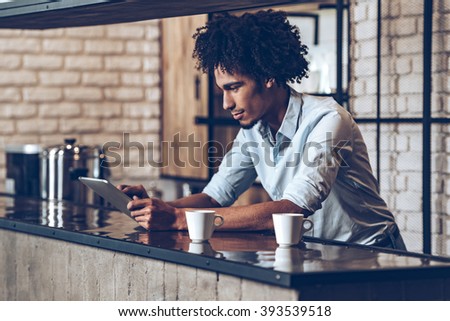 Running business easier with technologies. Side view of young African man using his digital tablet while leaning at bar counter with two coffee cups