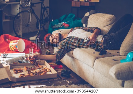 He had too much beer. Young handsome man passed out on sofa with pizza slice and beer can in his hand in messy room after party