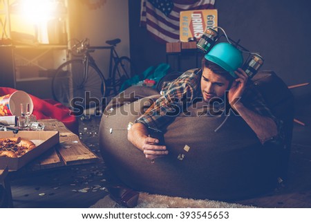 Need to delete that picture! Handsome young man in beer hat using his smart phone while lying on bean bag in messy room after party