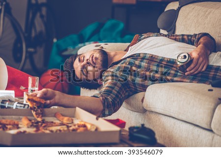 It has been a long night. Young handsome man passed out on sofa with pizza slice and beer can in his hand in messy room after party