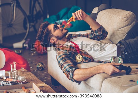 Still got energy to party. Young handsome man in sunglasses eating pizza and holding joystick in his hand while lying on sofa in messy room after party