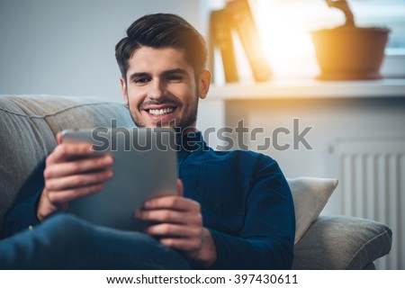 Spending great time at home. Close-up of handsome young man using his digital tablet with smile while lying down on the couch at home