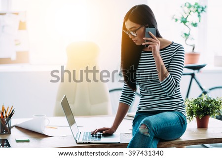Casual working day of architect. Beautiful young Asian woman in glasses talking on mobile phone and using laptop while sitting on the table at her working place