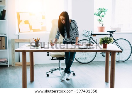 Working on new project. Full length of young beautiful Asian woman making some notes at blueprints while leaning to the table in office