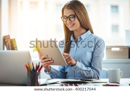 Lets check my timetable! Cheerful young beautiful woman in glasses using her touchpad with smile while sitting at her working place