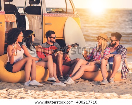 Carefree fun. Group of joyful young people drinking beer and playing guitar while sitting on the beach near their retro minivan