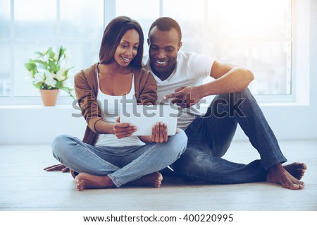Examining their new tab. Beautiful young African couple using digital tablet while sitting together on the floor at their apartments