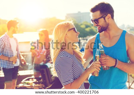 Cheers! Smiling young couple clinking glasses with beer and looking at each other while two people barbecuing in the background