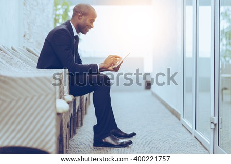 Checking his business schedule. Side view of cheerful young African businessman working on digital tablet while sitting on the chair