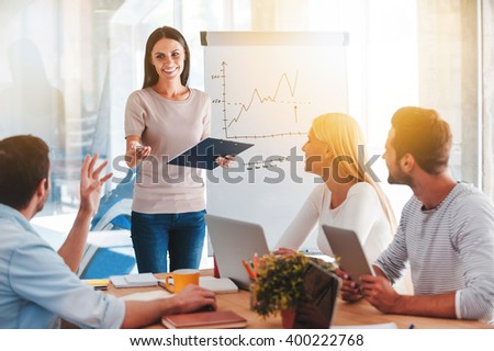 Discussing new business ideas. Cheerful young woman standing near whiteboard and smiling while her colleagues sitting at the desk
