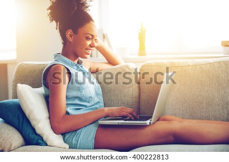 Surfing web at home. Side view of attractive young African woman working on laptop and smiling while sitting on the couch at home